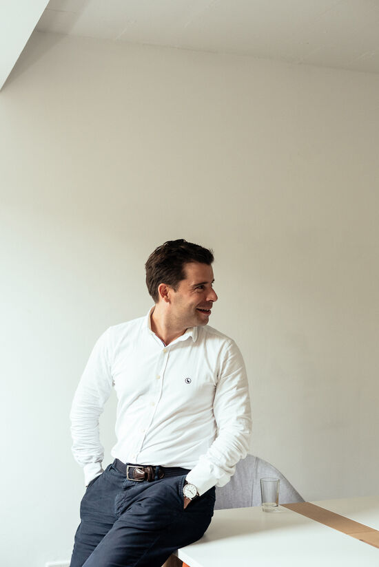 Felix Haas mit weißem Hemd und blauer Hose sitzt an der Tischkante und schaut zur Seite