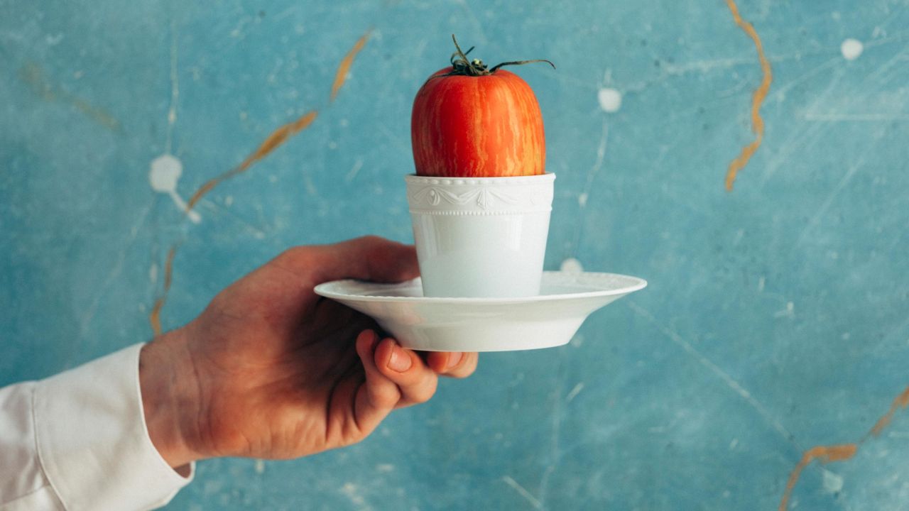 Eine Person hält eine Tasse mit einer Tomate in der Hand, im Hintergrund eine blau-braune Mamorwand. Eine Person hält eine Tasse mit einer Tomate in der Hand, im Hintergrund eine blau-braune Mamorwand.
