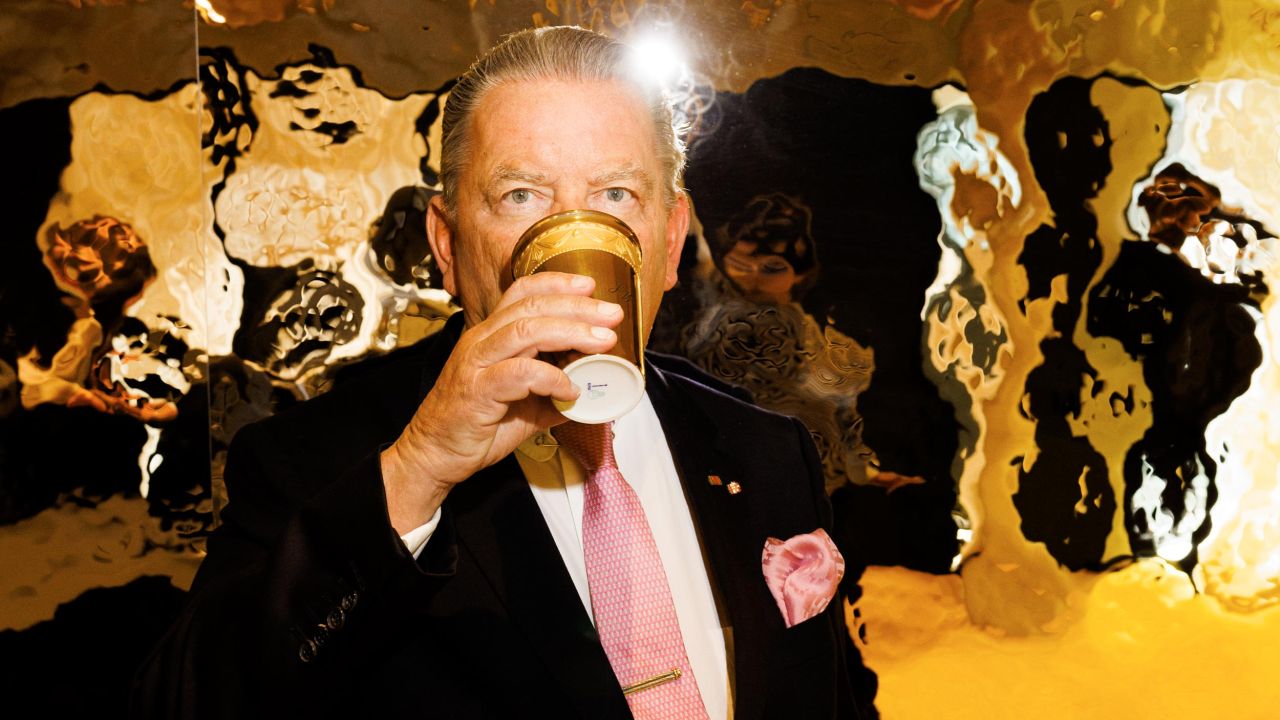 Nahaufnahme eines Mannes in Anzug und Krawatte, der aus einem goldenen Becher trinkt, mit einer stark reflektierenden goldenen Wand im Hintergrund Nahaufnahme eines Mannes in Anzug und Krawatte, der aus einem goldenen Becher trinkt, mit einer stark reflektierenden goldenen Wand im Hintergrund