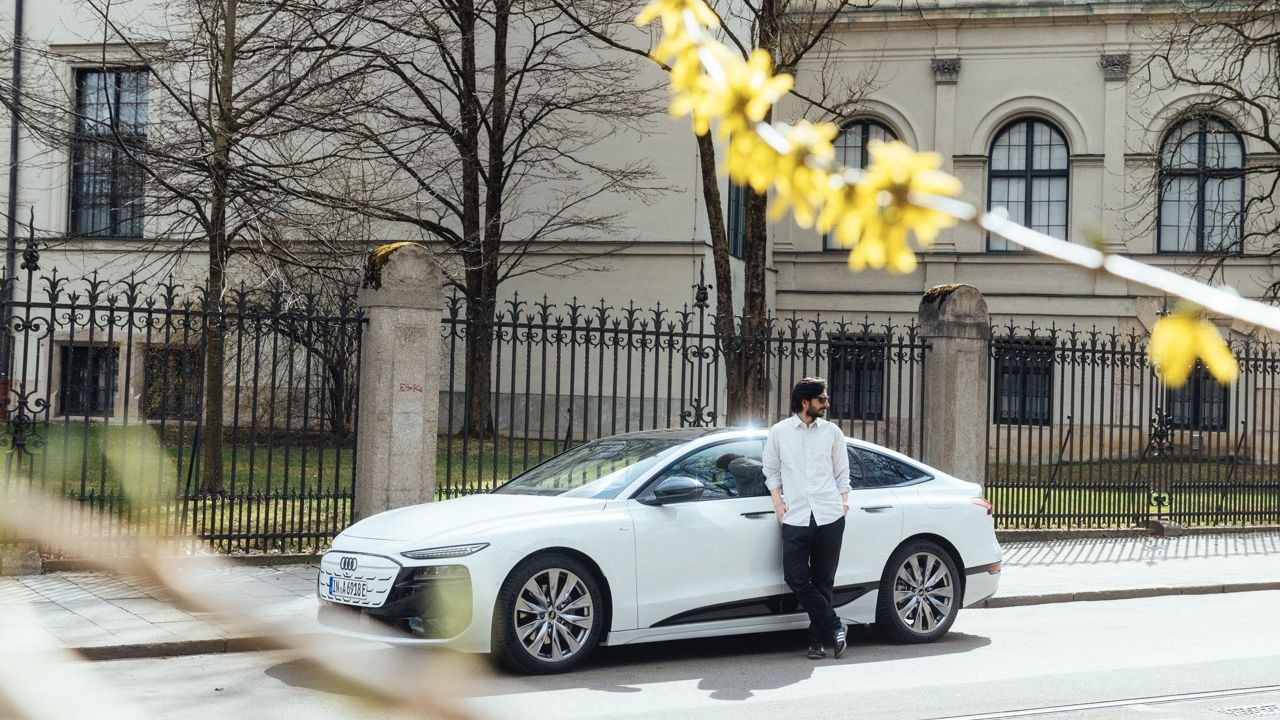 Ein Mann lehnt an der Seite eines weißen Audi A6 Sportback e-tron, der in einer städtischen Umgebung geparkt ist.