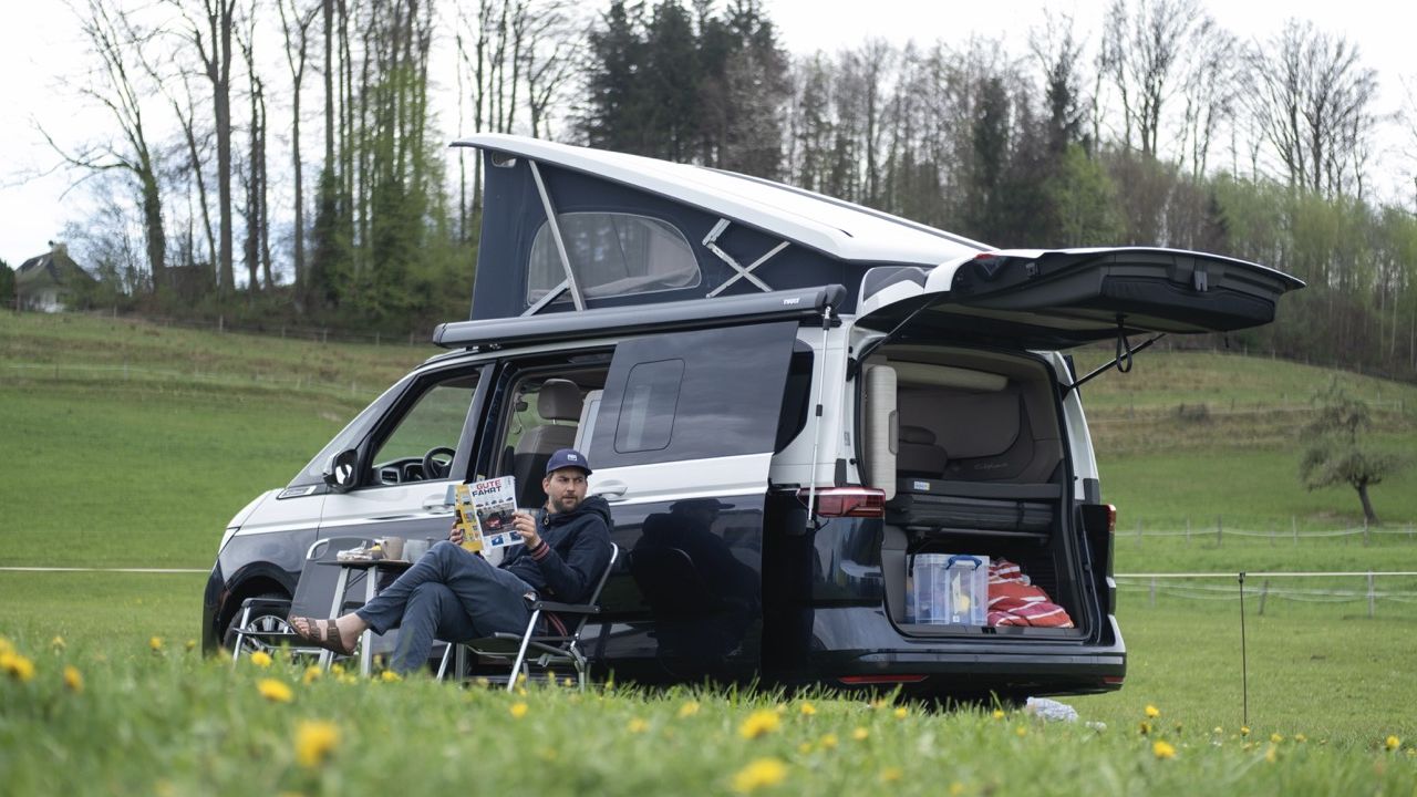 Ein Mann entspannt in einem Campingstuhl vor seinem VW California mit ausgefahrenem Aufstelldach und Markise