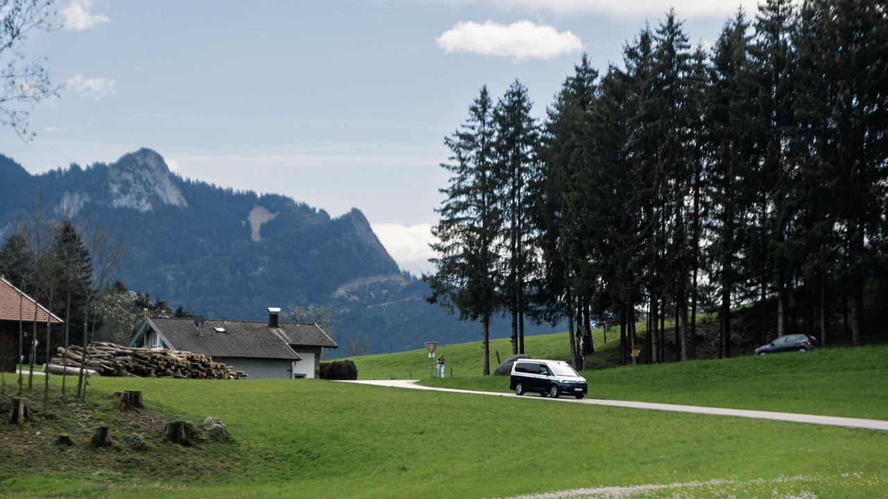 Dunkler VW California Camper fährt auf einer schmalen Straße durch eine grüne Hügellandschaft im Voralpenland