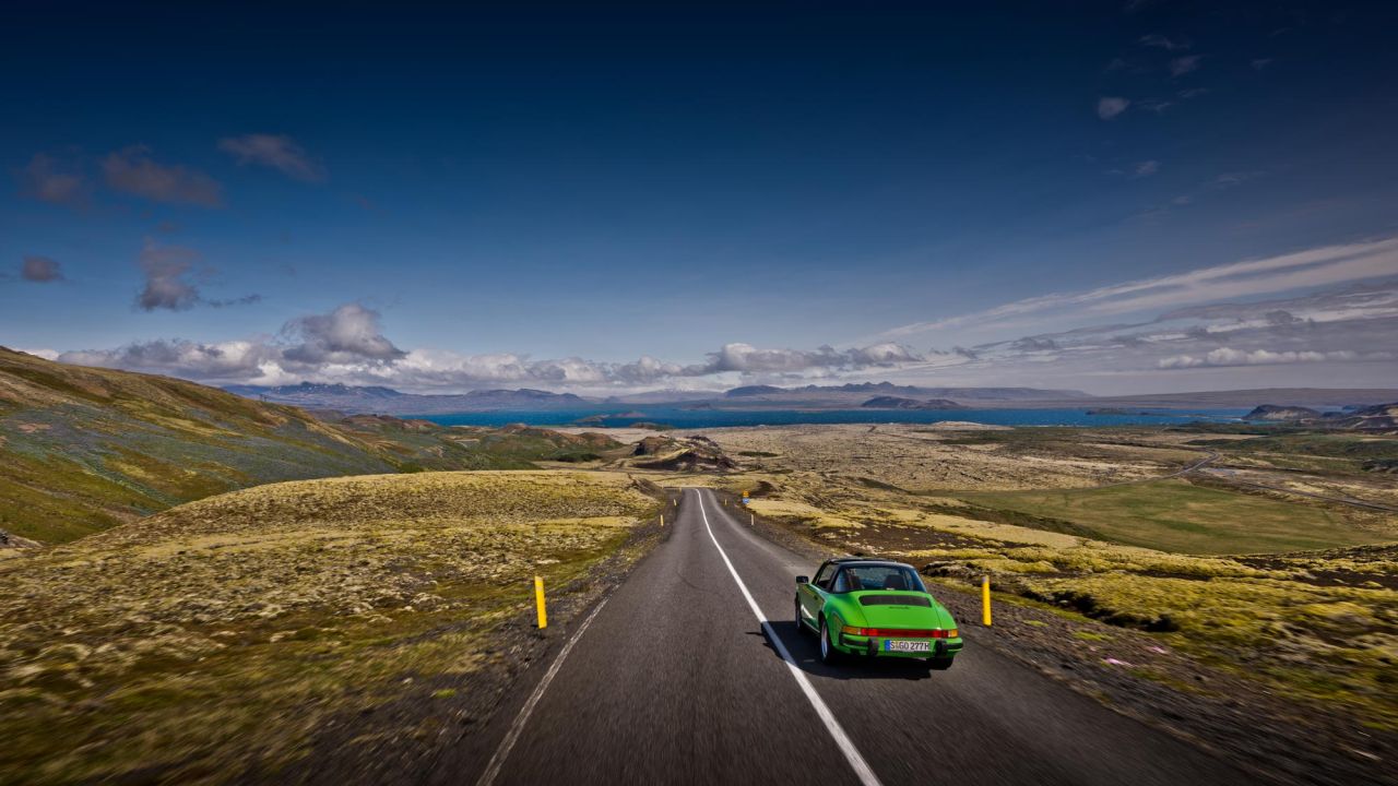 Ein grüner Porsche Oldtimer fährt über eine leere Landstraße durch eine bergige Landschaft in Island.