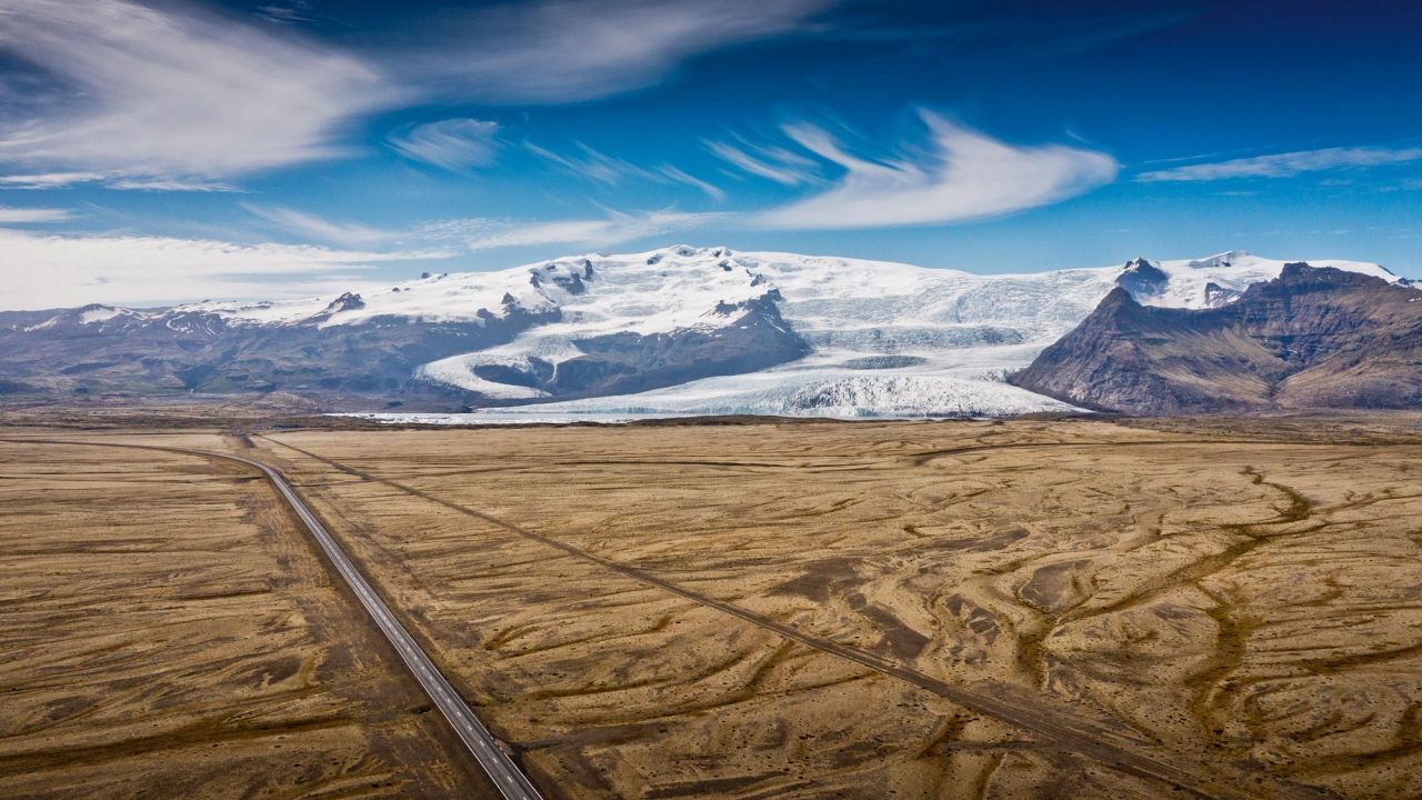 Luftaufnahme einer leeren Straße, die durch eine karge Ebene mit Gletscher und Bergen in Island führt.