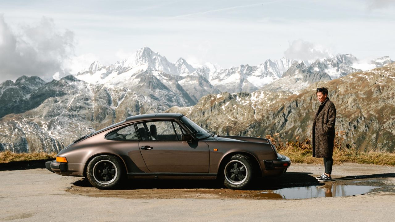 Ein brauner Porsche 911 steht auf einer Bergstraße vor einem Panorama schneebedeckter Alpengipfel, neben dem Auto steht eine Frau