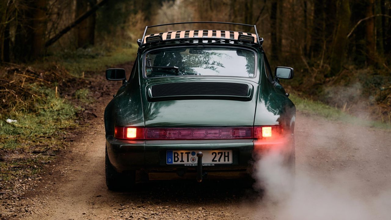 Heckansicht eines dunkelgrünen Porsche 911 mit Dachgepäckträger, der auf einem Waldweg fährt