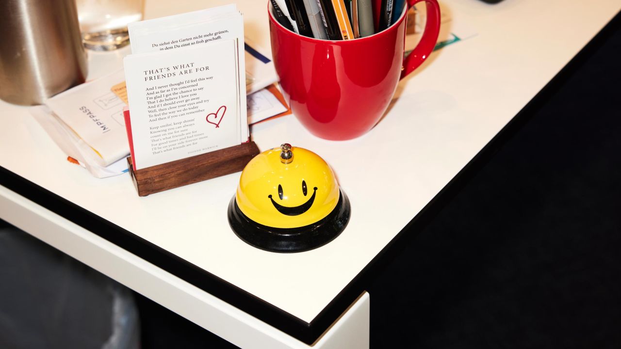 Gelb-schwarze Tischglocke mit einem Smiley steht auf einem weißen Schreibtisch neben einer Visitenkartenhalterung und einer roten Stiftekiste